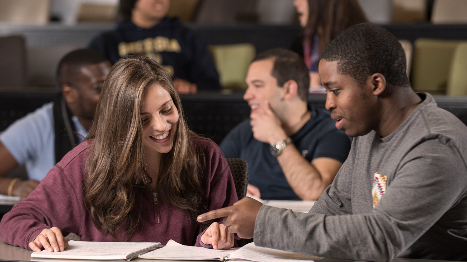 A young white woman and young Black man looking at a notebook in a college classroom.