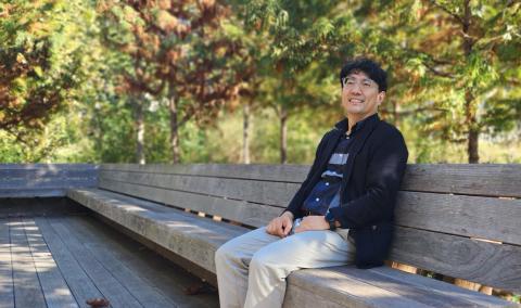 Sanghyun Jang seated on a wooden bench outdoors among trees.